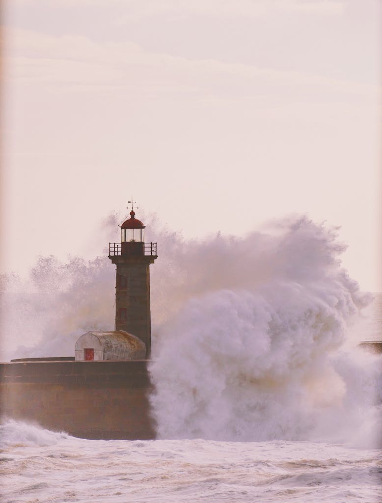 Powerful ocean waves crash against a solitary lighthouse at sunset, creating a dramatic and captivating scene.
